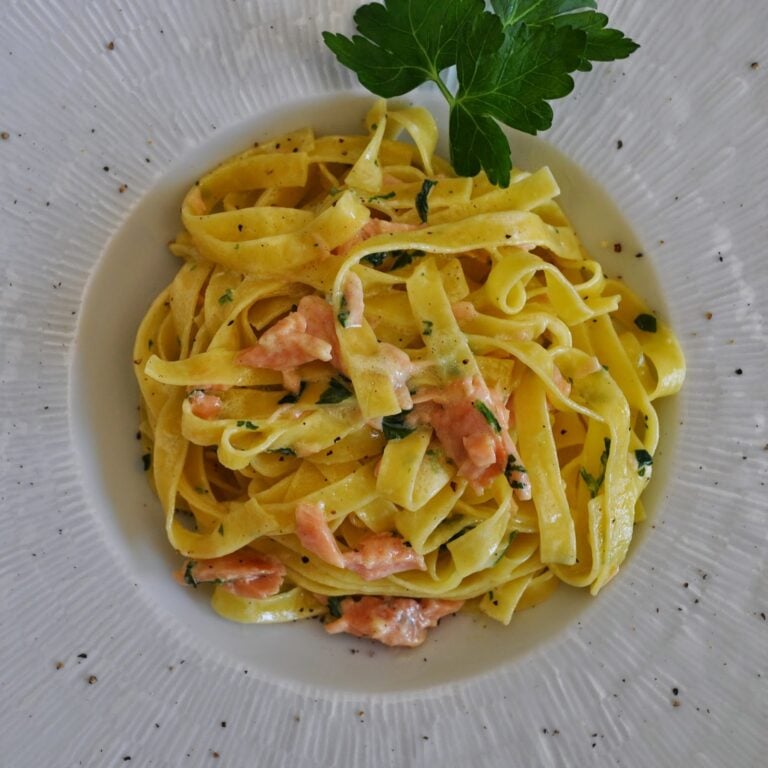 A plate of tagliatelle pasta topped with a creamy salmon sauce and garnished with fresh parsley, served on a white dish with a textured rim.