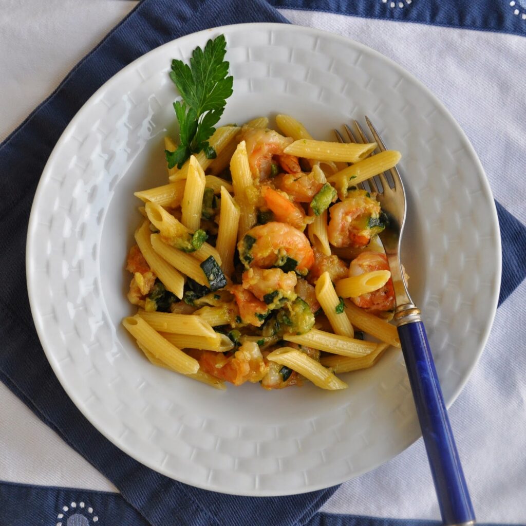 A white bowl filled with penne pasta, shrimp, and vegetables, garnished with a sprig of parsley. A fork rests on the side of the bowl, which is set on a blue and white cloth.