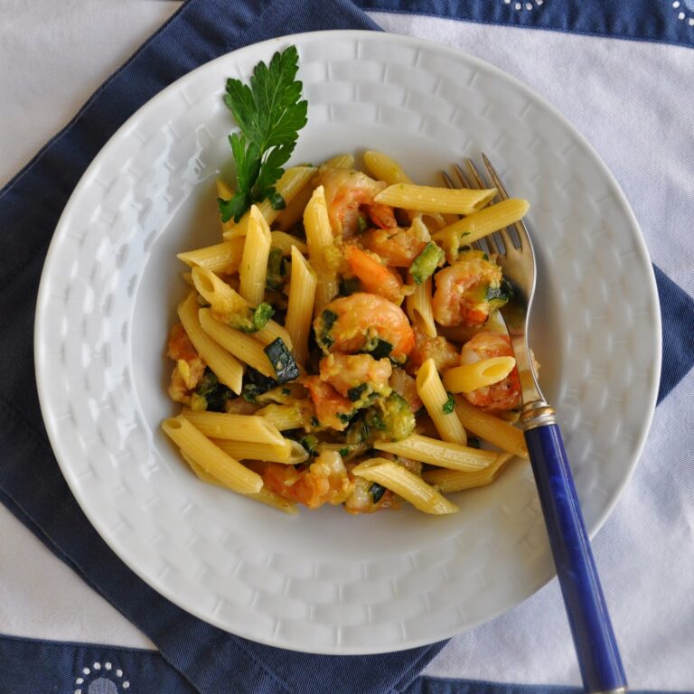 A white bowl filled with penne pasta, shrimp, and vegetables, garnished with a sprig of parsley. A fork rests on the side of the bowl, which is set on a blue and white cloth.