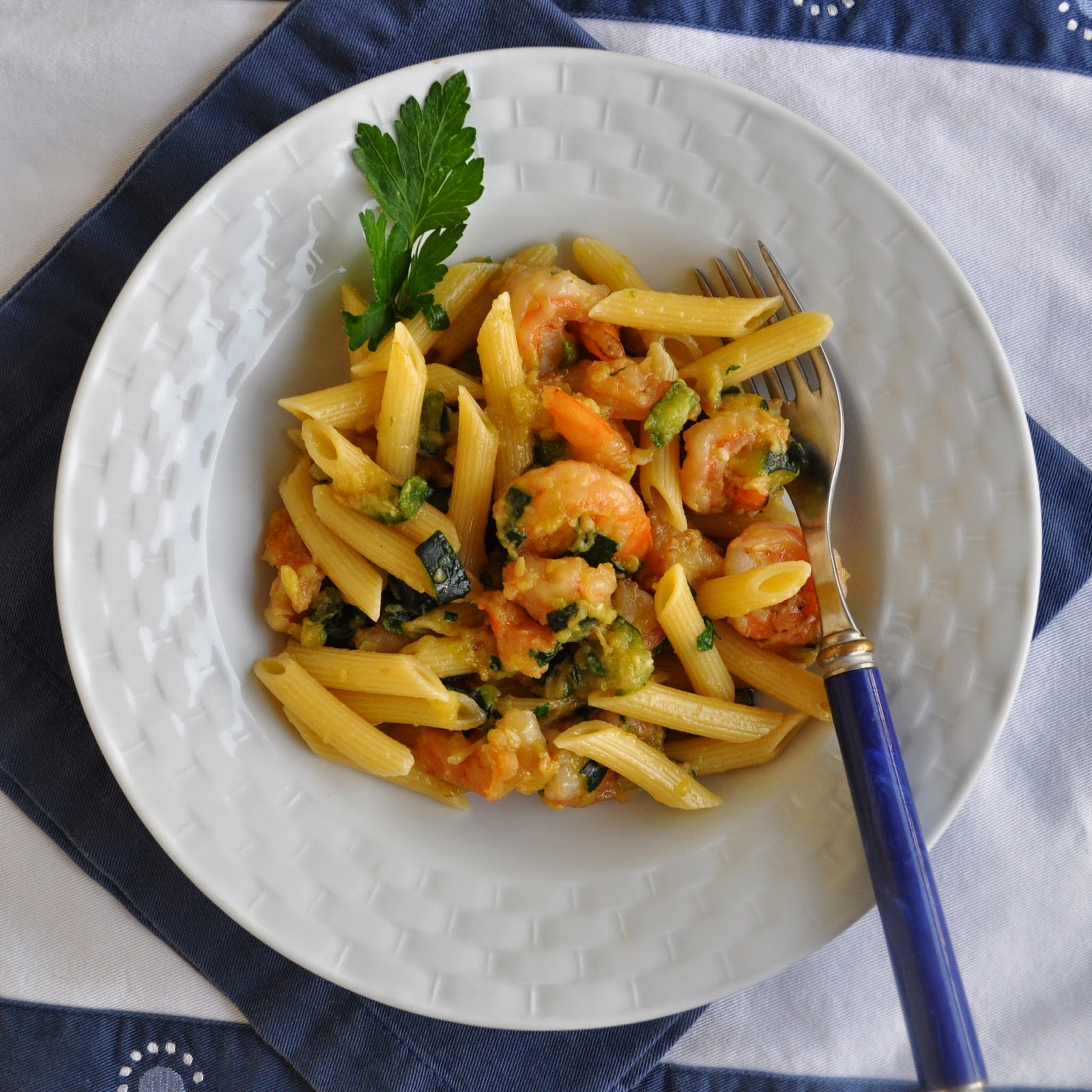 A white bowl filled with penne pasta, shrimp, and vegetables, garnished with a sprig of parsley. A fork rests on the side of the bowl, which is set on a blue and white cloth.