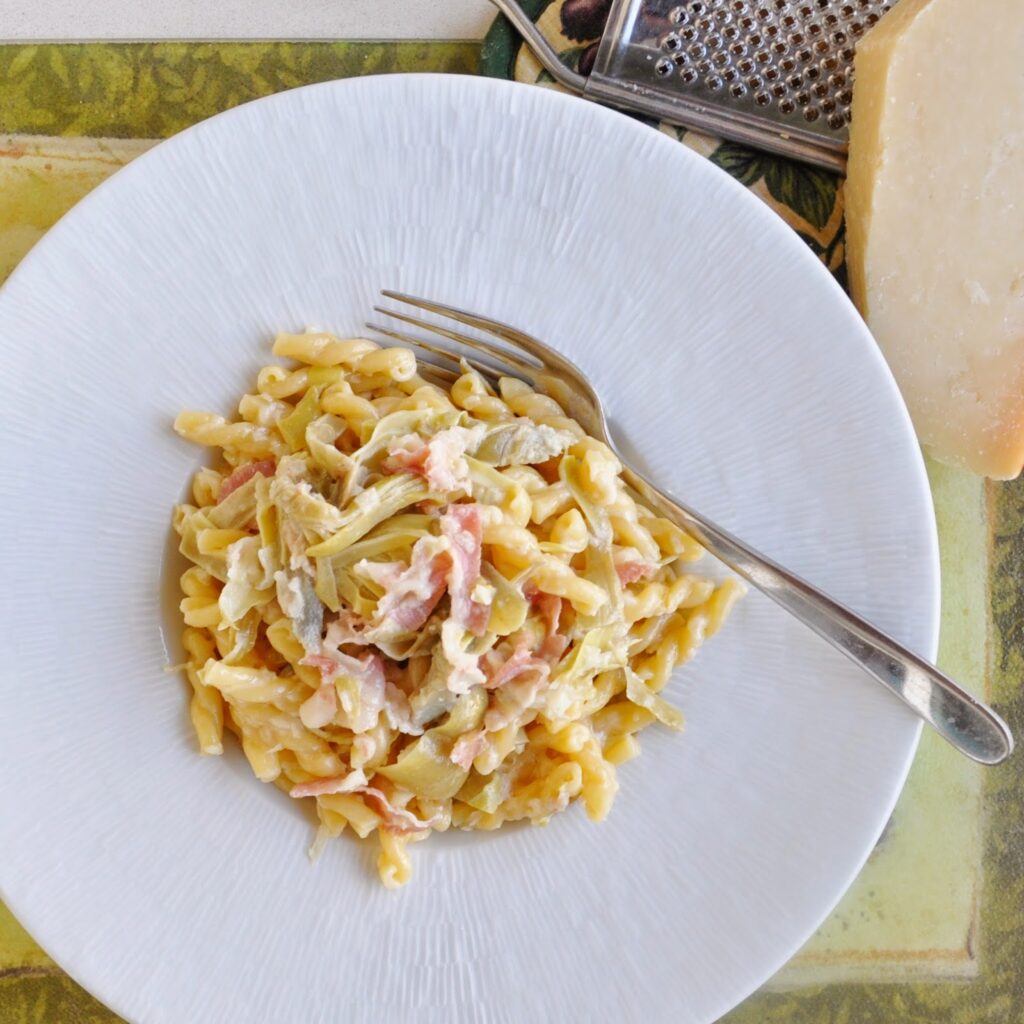 A plate of creamy pasta with strips of ham or bacon and grated cheese, served on a white dish with a fork. A cheese grater and a block of cheese are visible beside the plate.