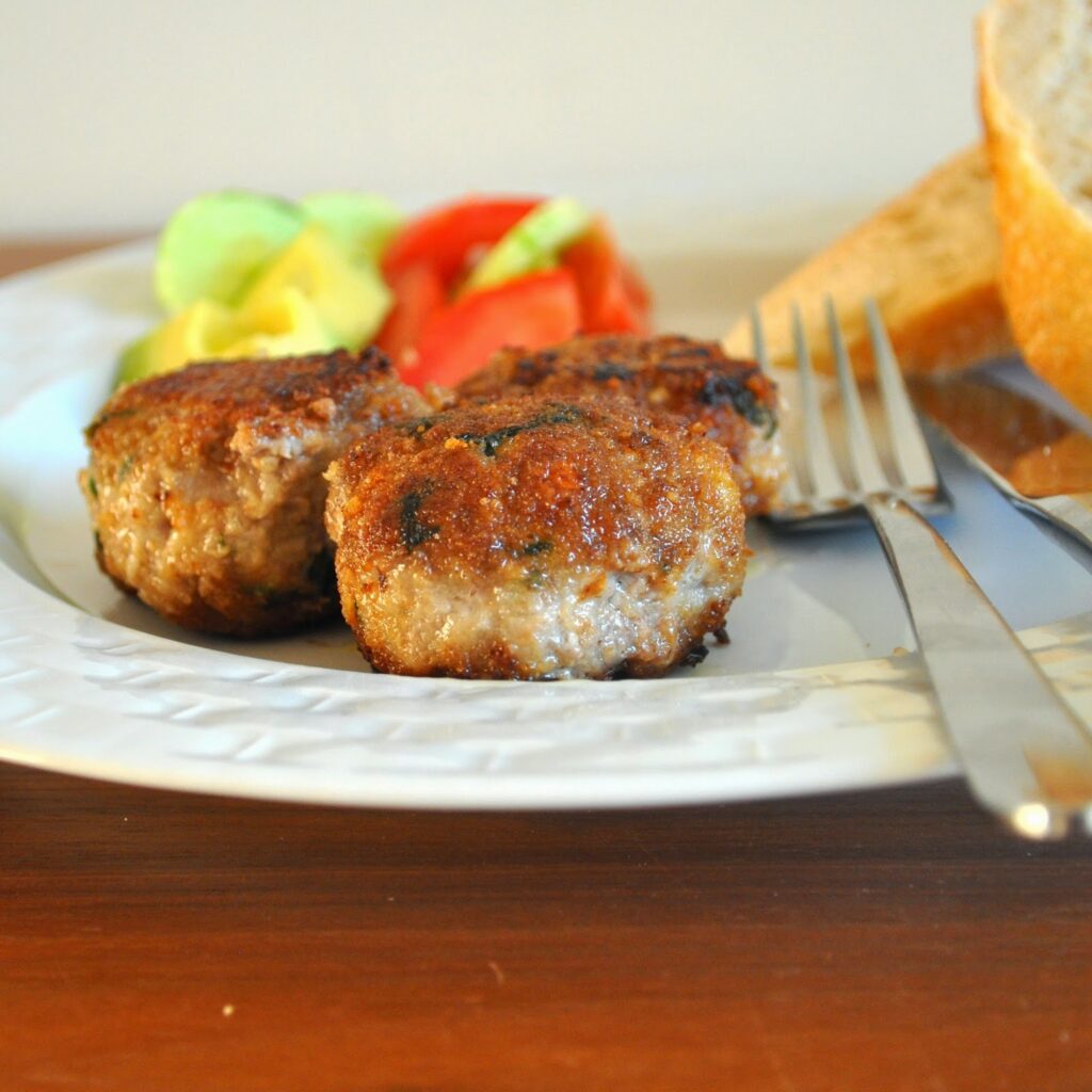 Three browned meat patties on a white plate with sliced tomato, lettuce, bread, and a fork and knife resting beside the food.