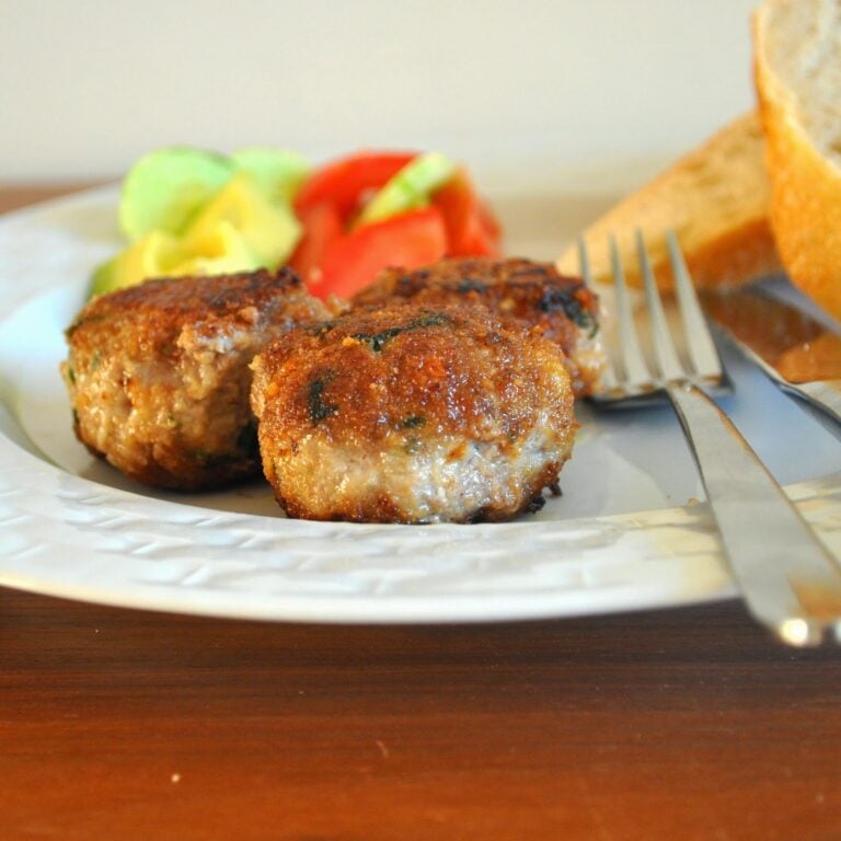 Three browned meat patties on a white plate with sliced tomato, lettuce, bread, and a fork and knife resting beside the food.