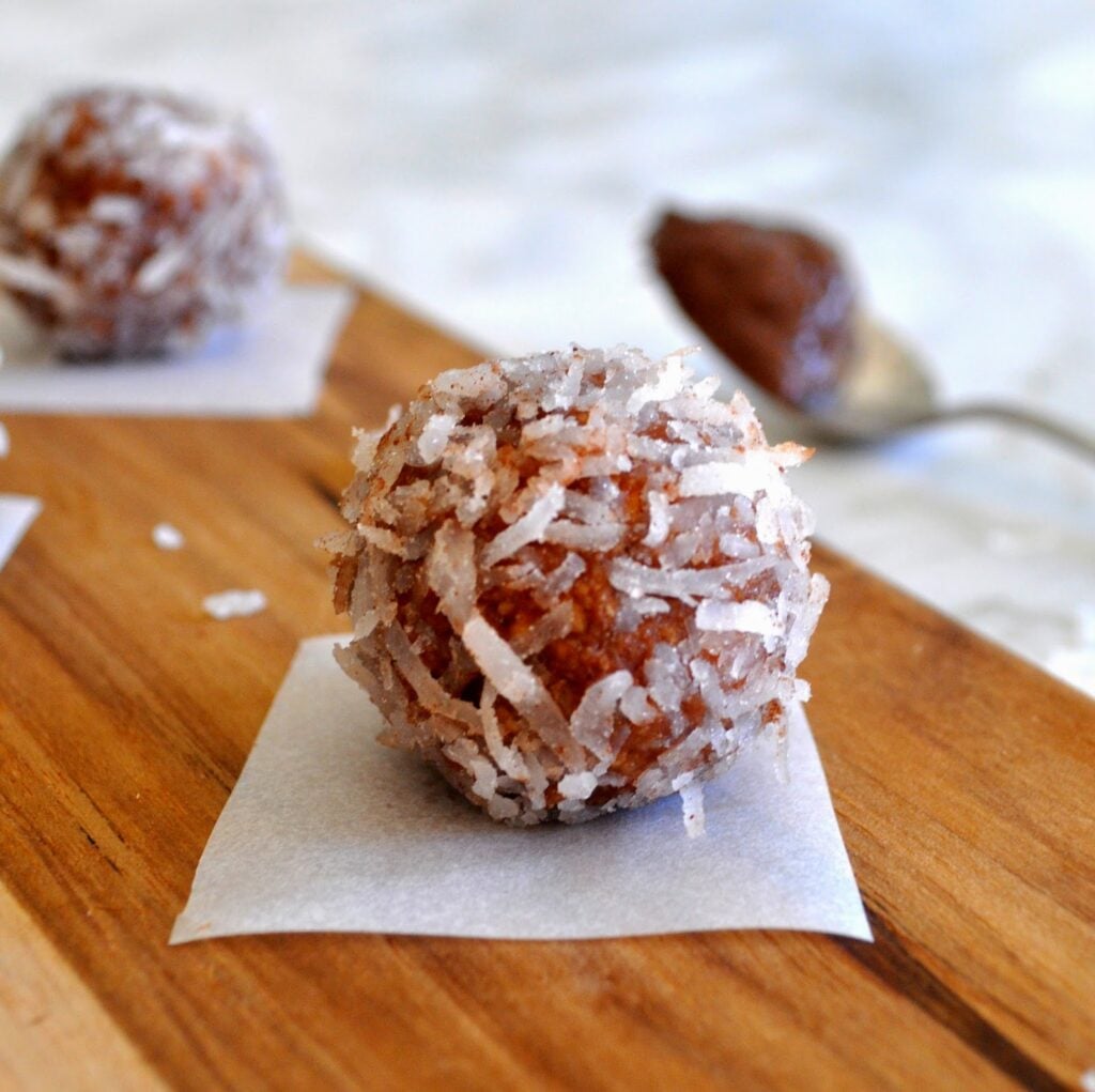A close-up of a chocolate ball coated in shredded coconut, placed on a small piece of parchment paper on a wooden surface, with a spoon and another treat blurred in the background.