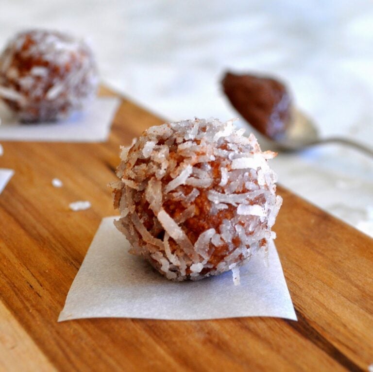 A close-up of a chocolate ball coated in shredded coconut, placed on a small piece of parchment paper on a wooden surface, with a spoon and another treat blurred in the background.
