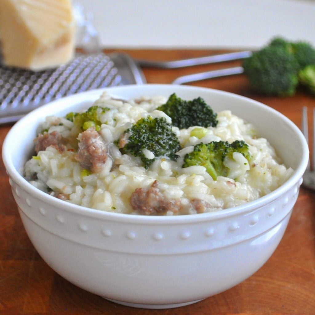 A white bowl filled with creamy risotto, featuring pieces of sausage and broccoli, sits on a wooden surface with a block of cheese and grater in the background.