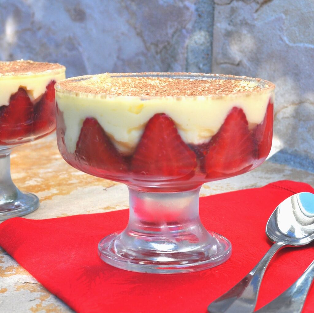 A glass dessert dish filled with layers of sliced strawberries, creamy custard, and a dusting of cocoa powder on top, placed on a red napkin beside a spoon and a stone background.