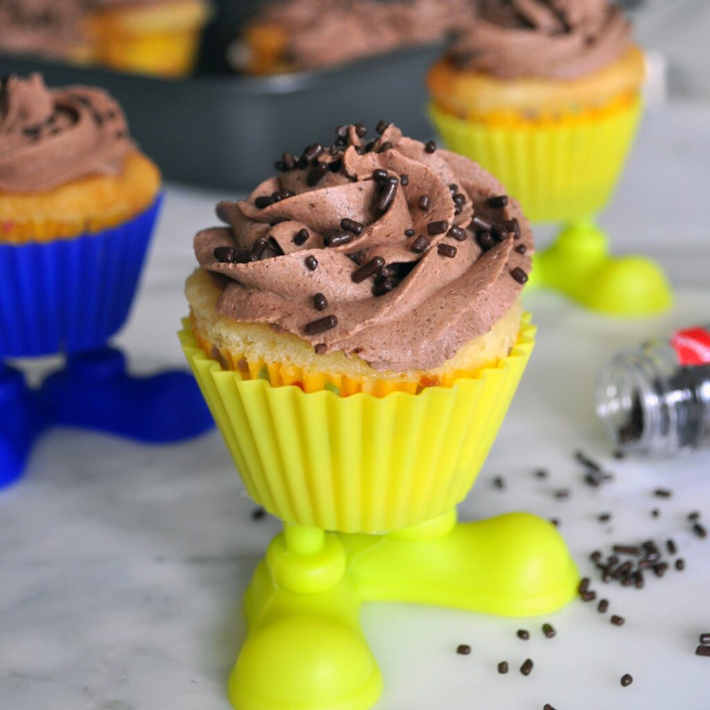 A cupcake in a yellow silicone holder with plastic feet stands on a marble surface. It is topped with chocolate frosting and sprinkles, with more cupcakes and sprinkles in the background.