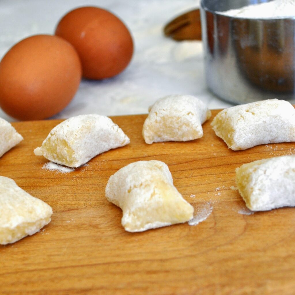 Pillow-shaped pieces of uncooked gnocchi dough dusted with flour on a wooden board, with two eggs and a metal measuring cup in the background.