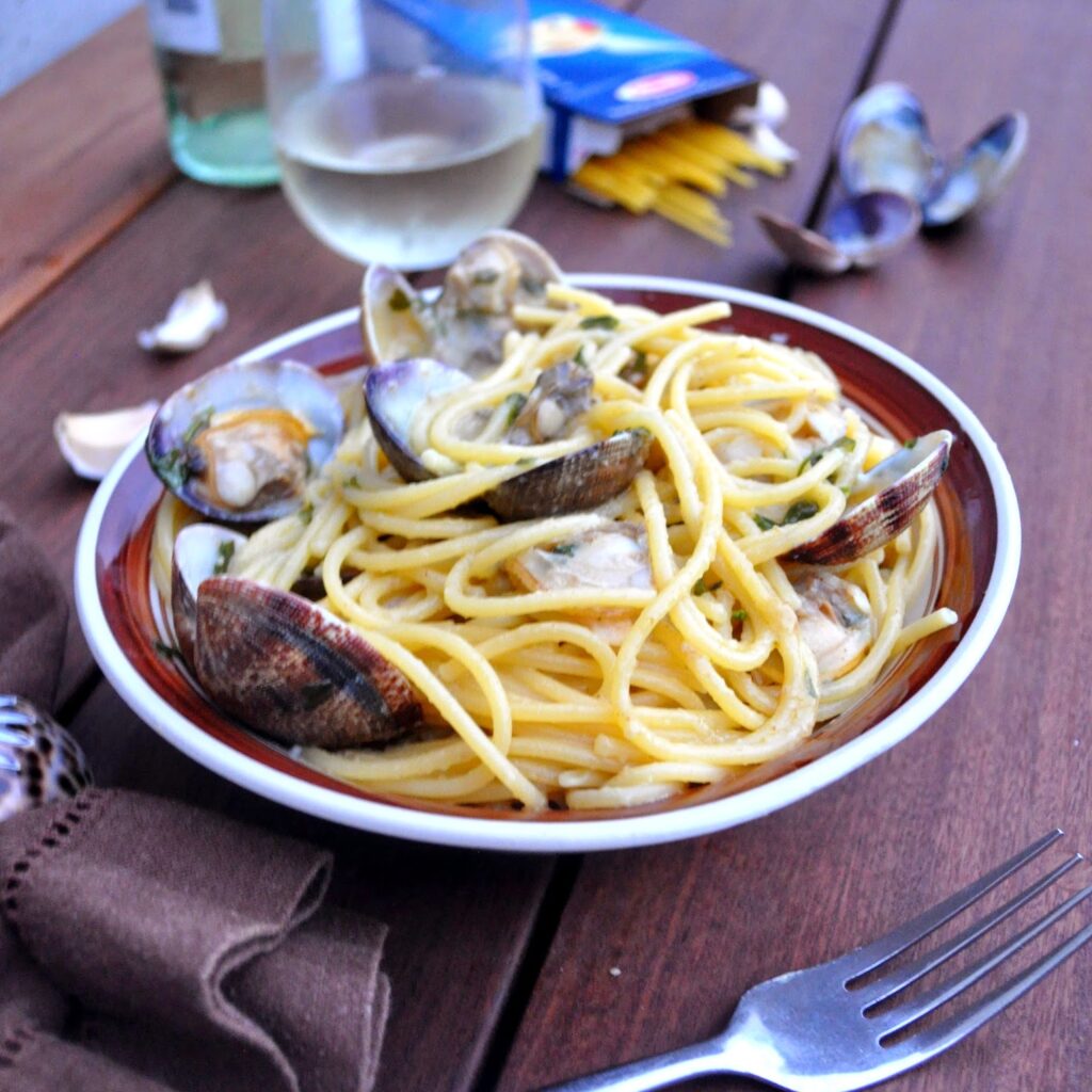 A bowl of spaghetti with clams in a creamy sauce sits on a wooden table. Nearby are a fork, a brown napkin, some garlic cloves, an open pasta box, and a glass of white wine.
