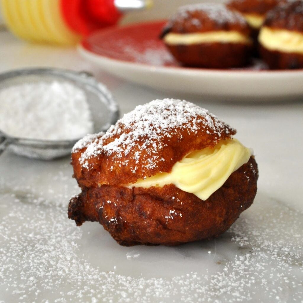 A golden-brown fried pastry filled with creamy yellow custard and dusted with powdered sugar, sitting on a marble surface with a sifter and more pastries on a plate in the background.