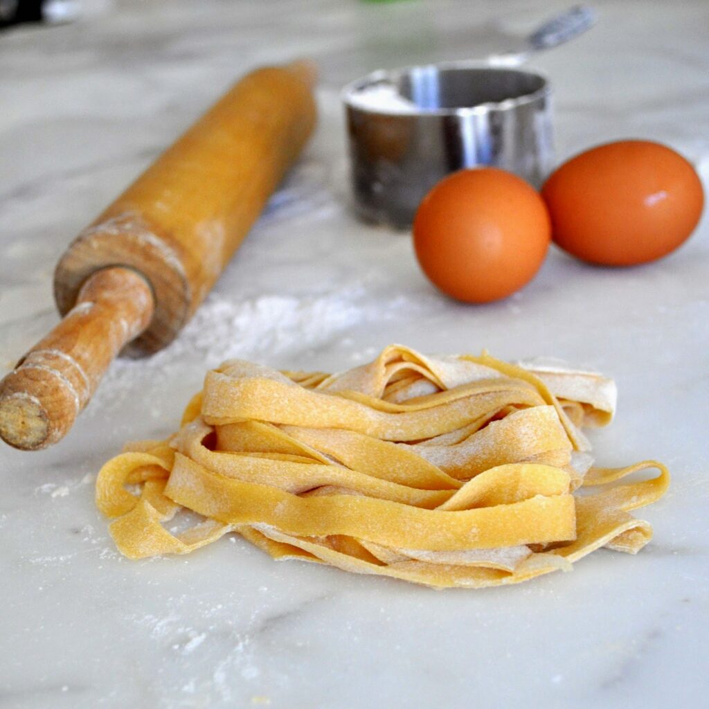 Freshly made pasta noodles sit on a floured marble surface next to a wooden rolling pin, two brown eggs, and a metal measuring cup in the background.