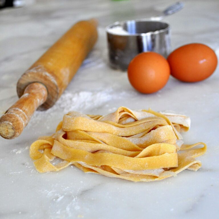 Freshly made pasta noodles sit on a floured marble surface next to a wooden rolling pin, two brown eggs, and a metal measuring cup in the background.