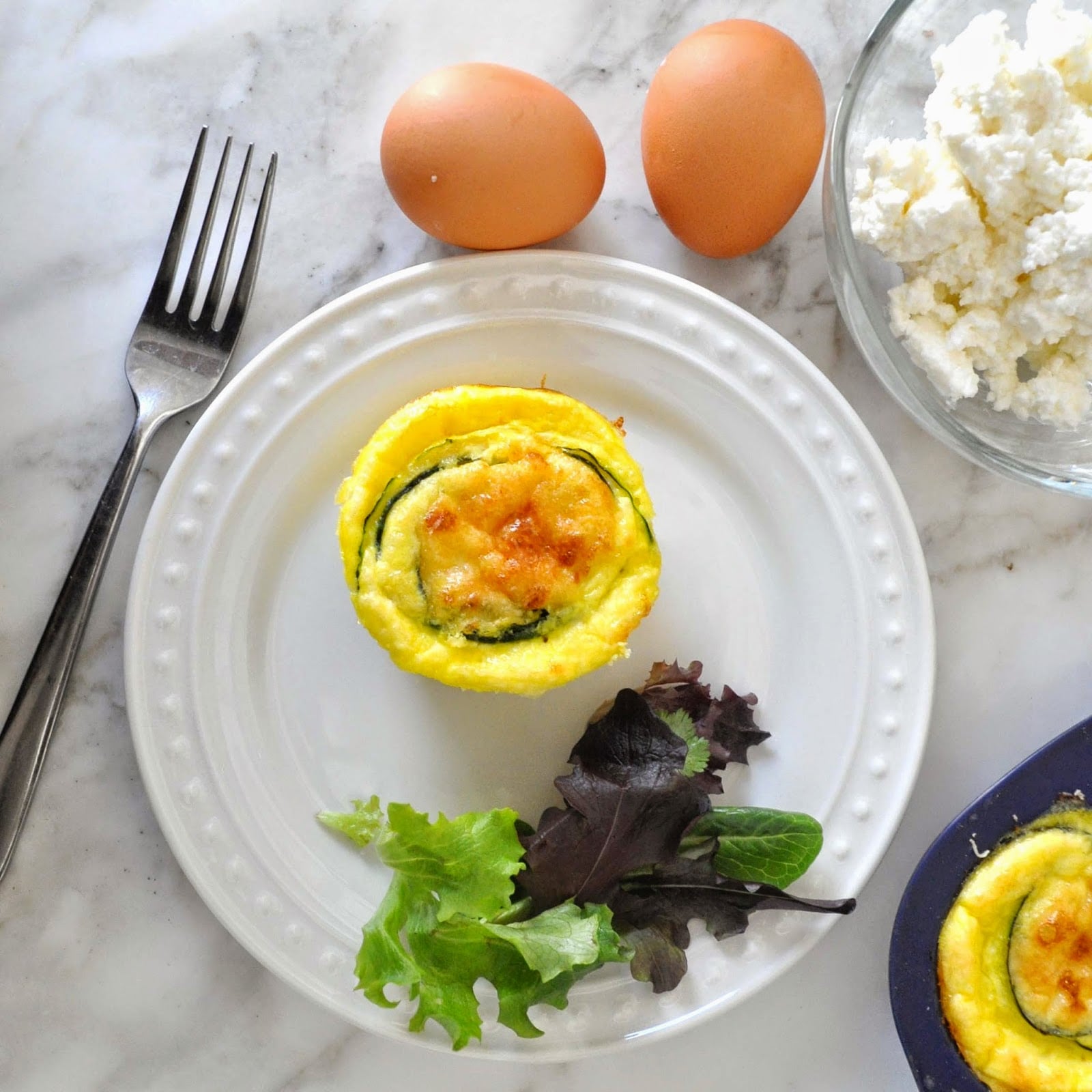 A mini quiche with zucchini spirals sits on a white plate with mixed greens. Nearby are two eggs, a bowl of cottage cheese, a fork, and part of a muffin tin with more quiches, all on a marble surface.