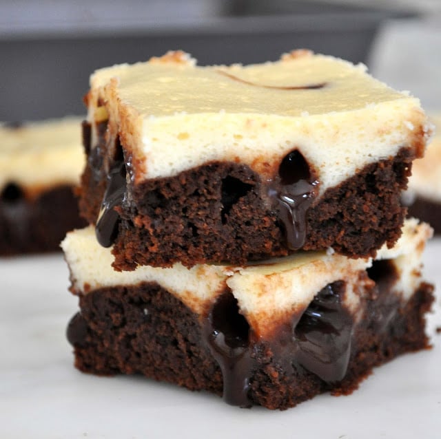 Two stacked cheesecake brownies with a fudgy chocolate center oozing out, featuring a creamy cheesecake layer on top and dense chocolate brownie on the bottom, set on a white surface.