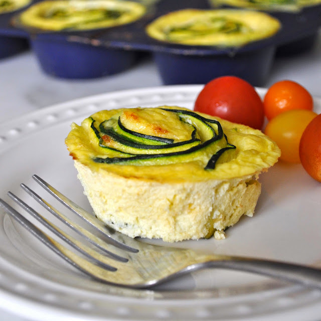 A mini zucchini quiche topped with spiraled zucchini slices sits on a white plate beside red and yellow cherry tomatoes, with a fork resting on the plate. More quiches are in the background.