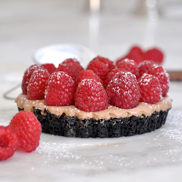 A chocolate tart topped with fresh raspberries sits on a marble surface, dusted with powdered sugar. A few loose raspberries are placed nearby.