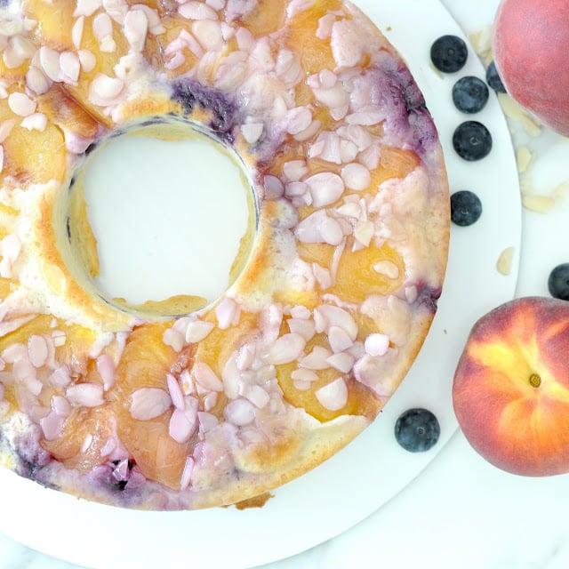 A ring-shaped upside-down cake topped with peach slices, blueberries, and almond slivers, displayed on a white surface with fresh peaches and blueberries nearby.