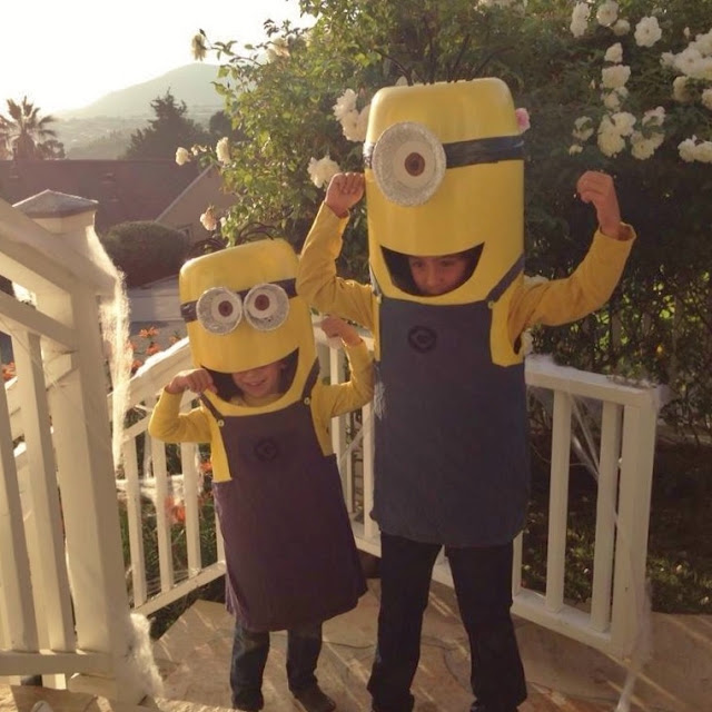 Two children dressed in homemade Minion costumes stand on a porch, flexing their arms in a playful pose. They wear yellow shirts, blue overalls, and helmets resembling Minion faces with large eyes.