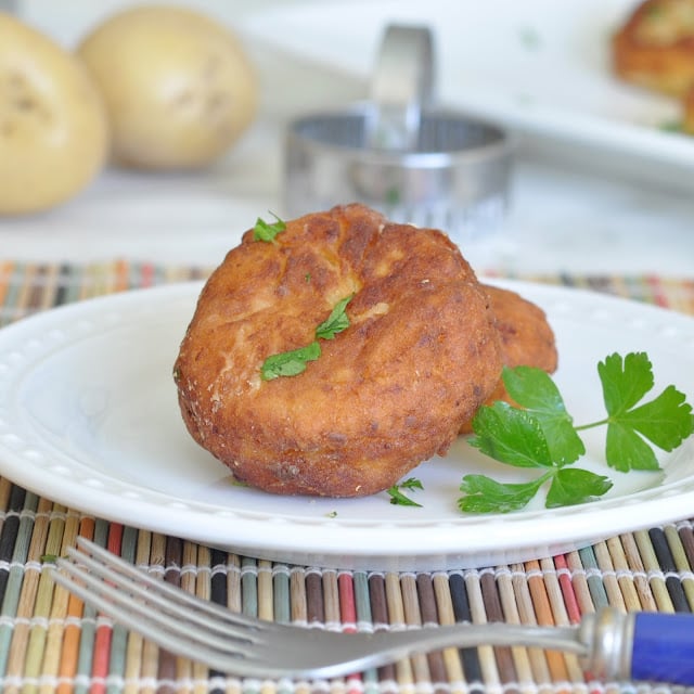 A golden-brown fritter garnished with fresh parsley sits on a white plate, with a fork nearby. Out-of-focus potatoes and a metal mold are visible in the background.