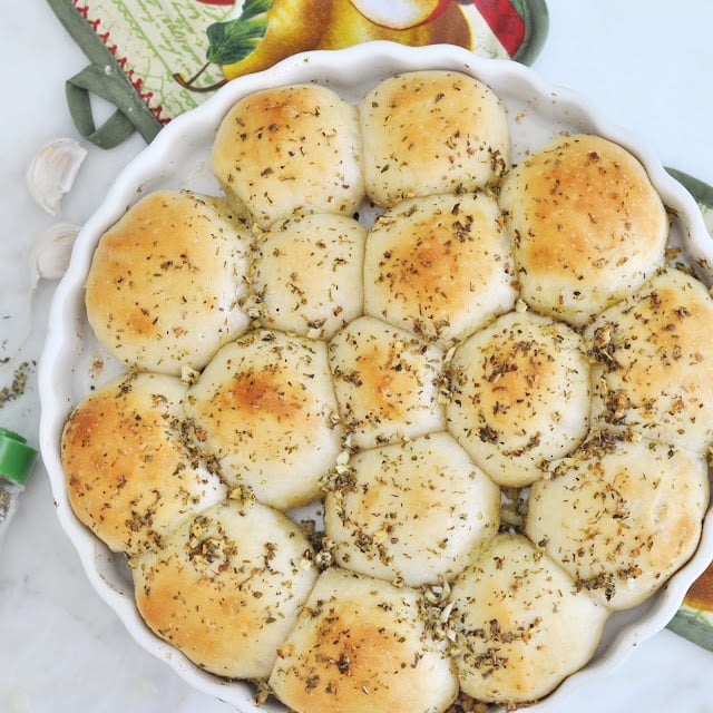 A round baking dish filled with golden-brown, pull-apart garlic herb rolls sprinkled with dried herbs, sitting on a marble surface next to a patterned oven mitt.