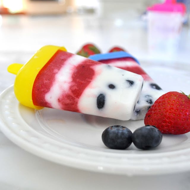 Homemade popsicles with red, white, and blue layers rest on a white plate, accompanied by fresh strawberries and blueberries on the side. The popsicles contain visible fruit pieces and are in colorful molds.