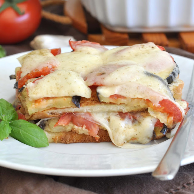 A close-up of a cheesy vegetable casserole slice on a white plate, with layers of bread, tomatoes, eggplant, and melted cheese. A sprig of basil and a tomato are visible in the background.