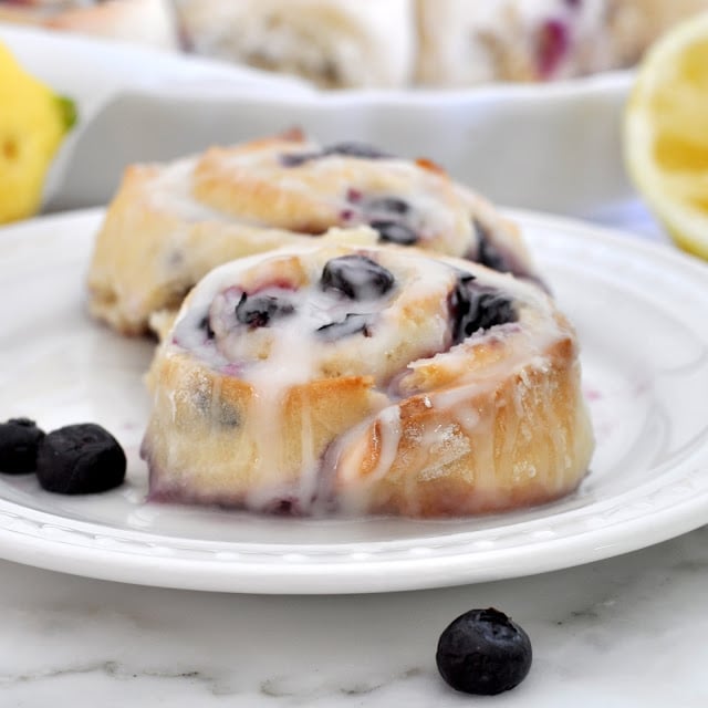 Two blueberry sweet rolls topped with icing sit on a white plate, with a few fresh blueberries nearby. The background includes a partially visible lemon and a baking dish.