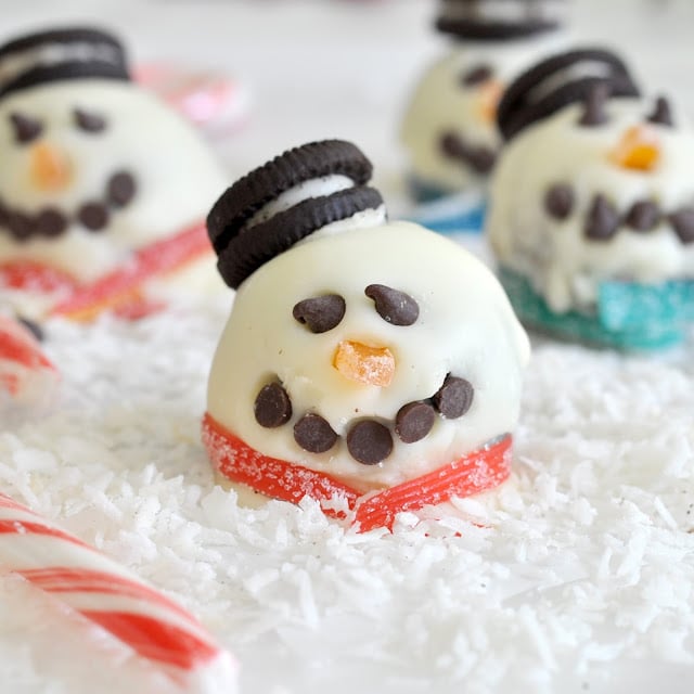 A close-up of a snowman-shaped treat made of white chocolate, Oreo cookies for a hat, candy for a scarf, chocolate chips for eyes and mouth, and an orange candy for the nose, with candy canes and shredded coconut “snow.”.