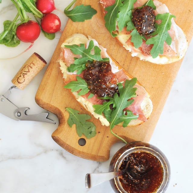 Open-faced sandwiches with prosciutto, arugula, and fig jam on a wooden cutting board, next to fresh radishes, a corkscrew, a wine cork, and a small bowl of fig jam on a white surface.