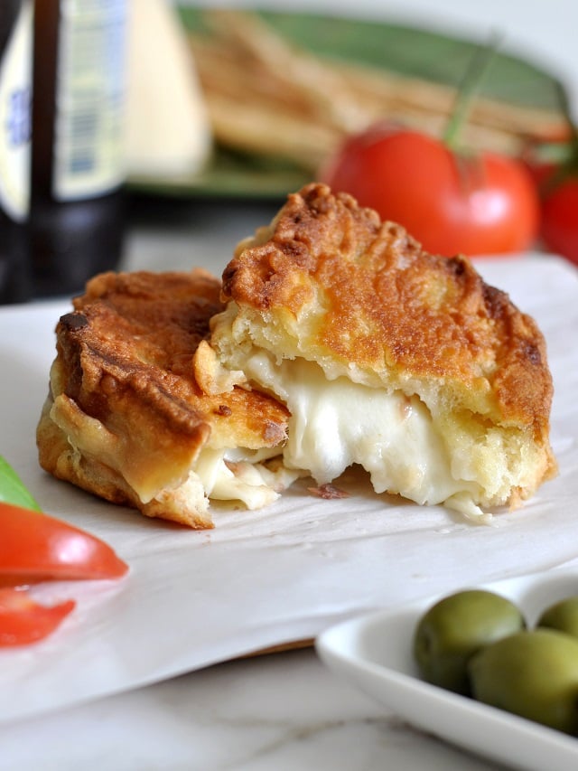 Two pieces of golden, crispy fried cheese sandwiches are placed on parchment paper, with melted cheese oozing out. Fresh tomatoes and green olives are nearby, with a blurred background of other ingredients.