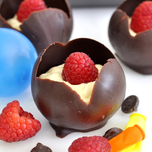 A chocolate sphere with a cracked top is filled with creamy custard and topped with a fresh raspberry. More raspberries, chocolate chips, a blue plastic mold, and a yellow object are nearby on a white surface.