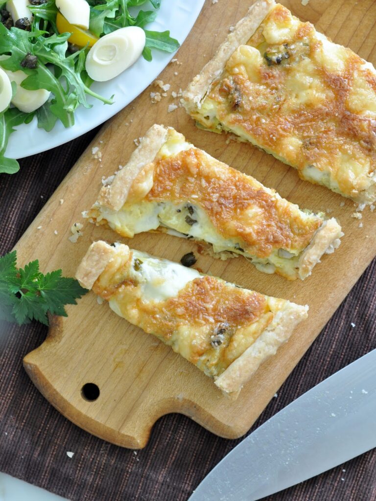 Three slices of cheesy tart are arranged on a wooden cutting board next to a large knife. A plate with leafy green salad and sliced vegetables is partially visible at the top left.
