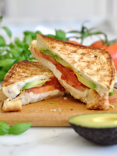 A grilled sandwich cut in half, filled with slices of tomato, avocado, and mozzarella cheese, sits on a wooden board. Fresh basil leaves and a halved avocado are nearby.