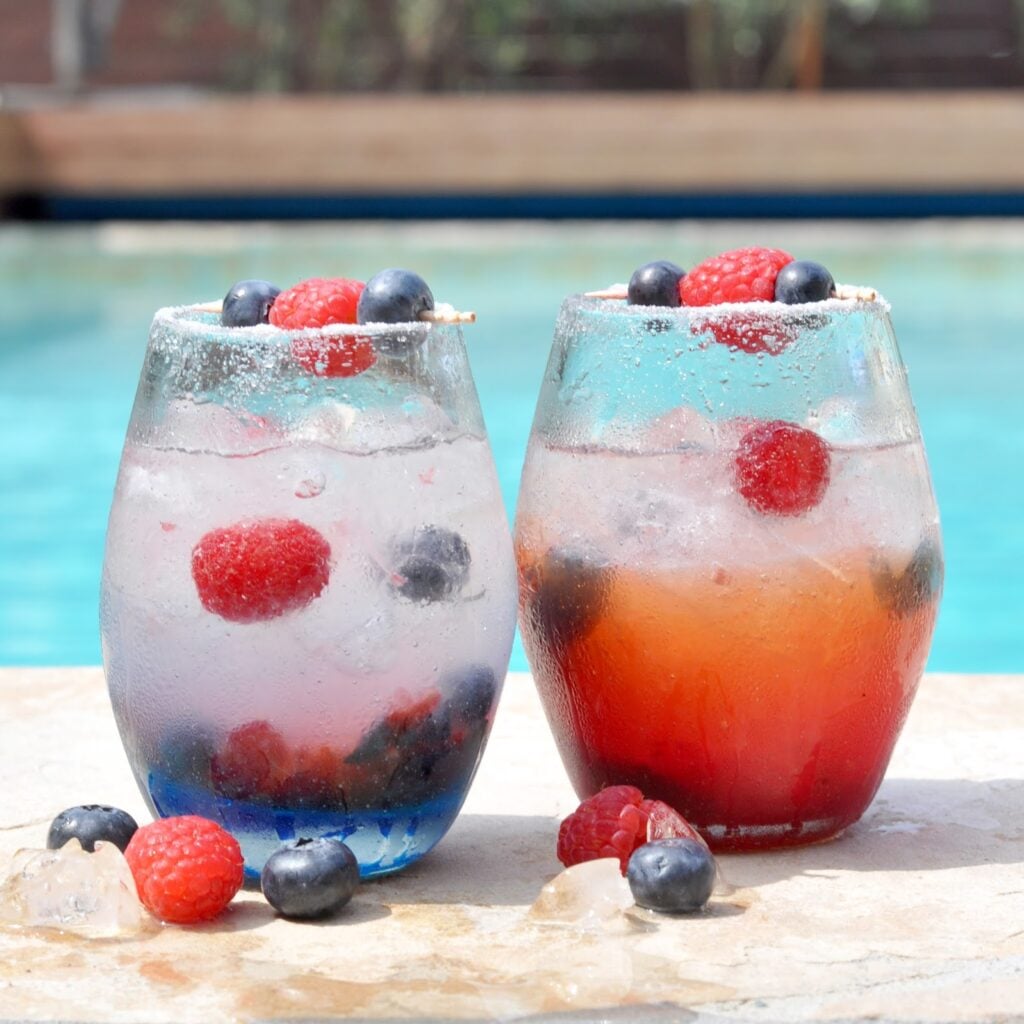 Two glasses of iced berry drinks sit by a pool, each garnished with blueberries and raspberries. Blueberries and raspberries are also scattered on the stone surface in front of the glasses.