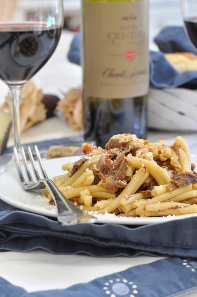 A plate of pasta with shredded meat and grated cheese, accompanied by a fork, is set on a blue napkin. Two glasses of red wine and a bottle of wine are in the background, creating a cozy dining atmosphere.
