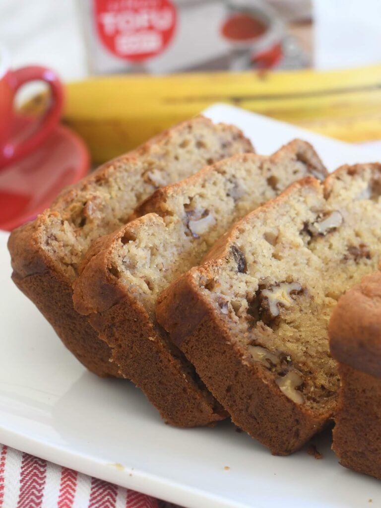 Three thick slices of banana nut bread are arranged on a white plate. The bread is golden brown with visible pieces of walnut inside. A banana and a red mug are blurred in the background.