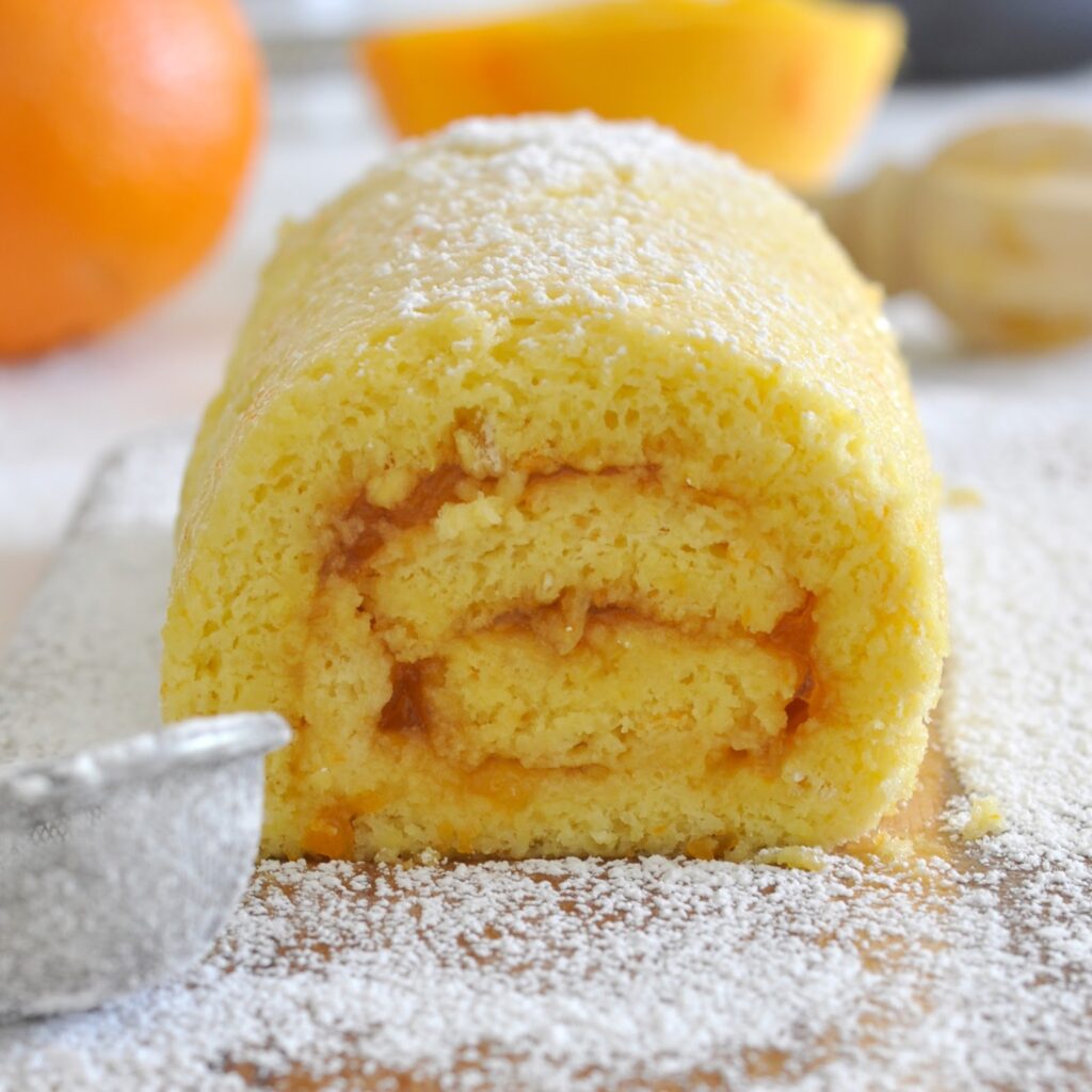 A close-up of a rolled sponge cake dusted with powdered sugar, showing its golden-yellow layers and swirl of filling inside. Blurred oranges and baking utensils are visible in the background.
