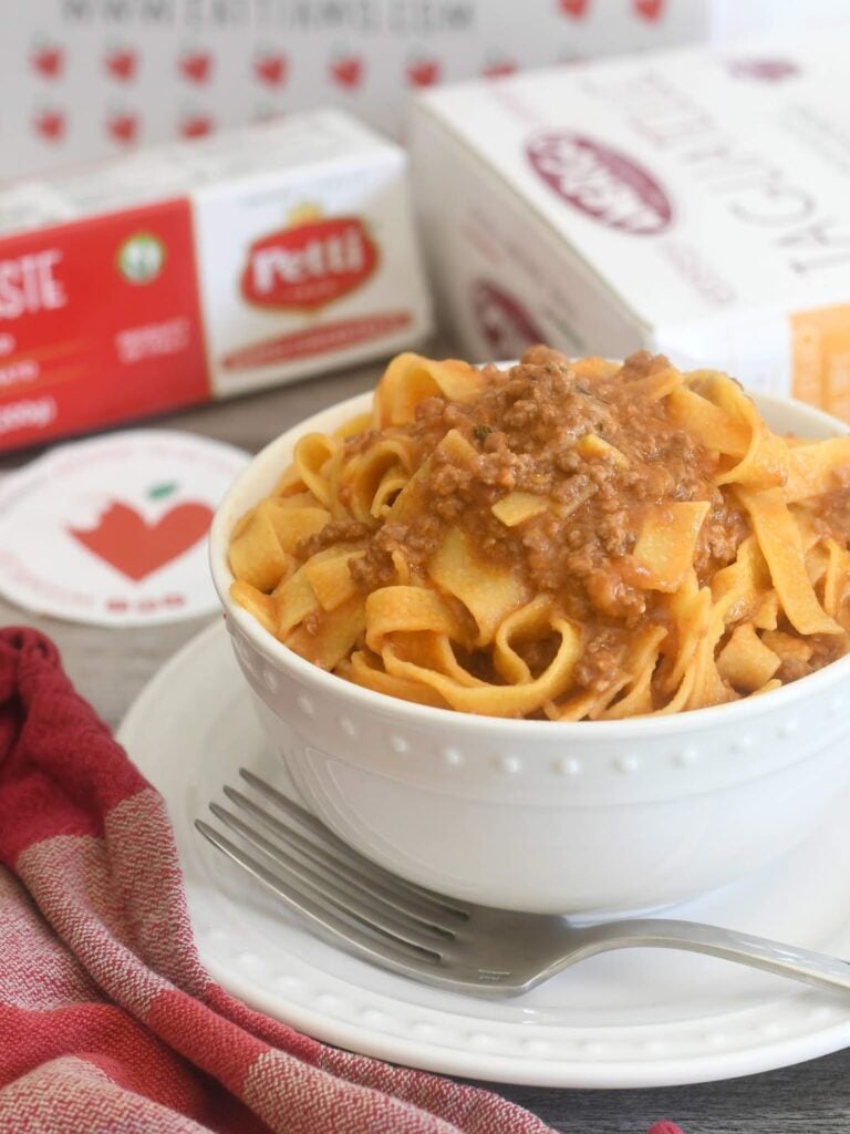 A white bowl of tagliatelle pasta topped with meat sauce sits on a white plate with a fork beside it. A red napkin is nearby, and boxes of tomato products are in the blurred background.