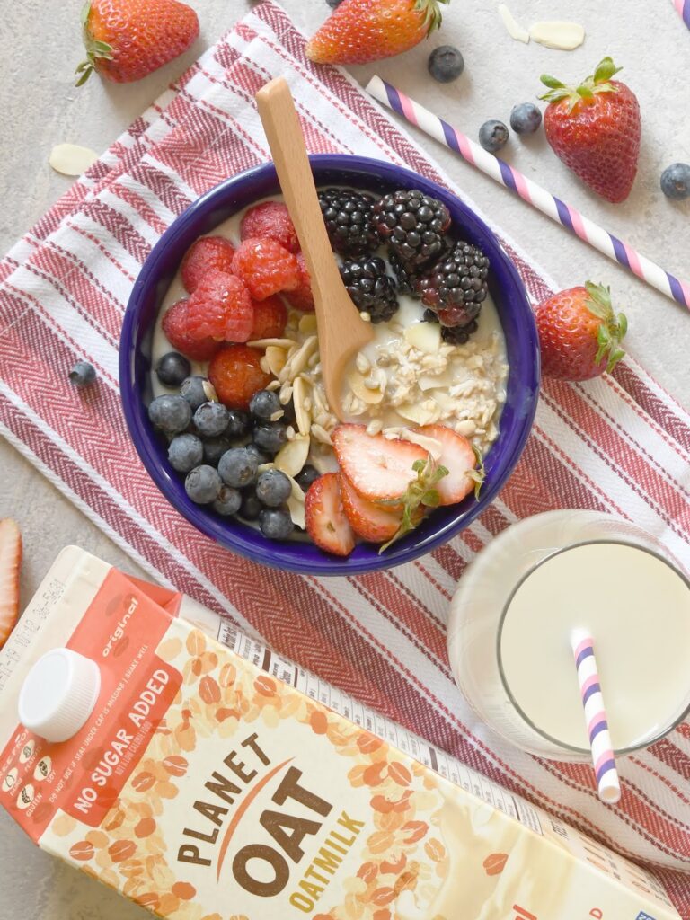 A bowl of oatmeal topped with strawberries, raspberries, blackberries, blueberries, and almond slices sits on a red-striped cloth, next to oat milk in a glass and carton, with scattered berries and a straw.
