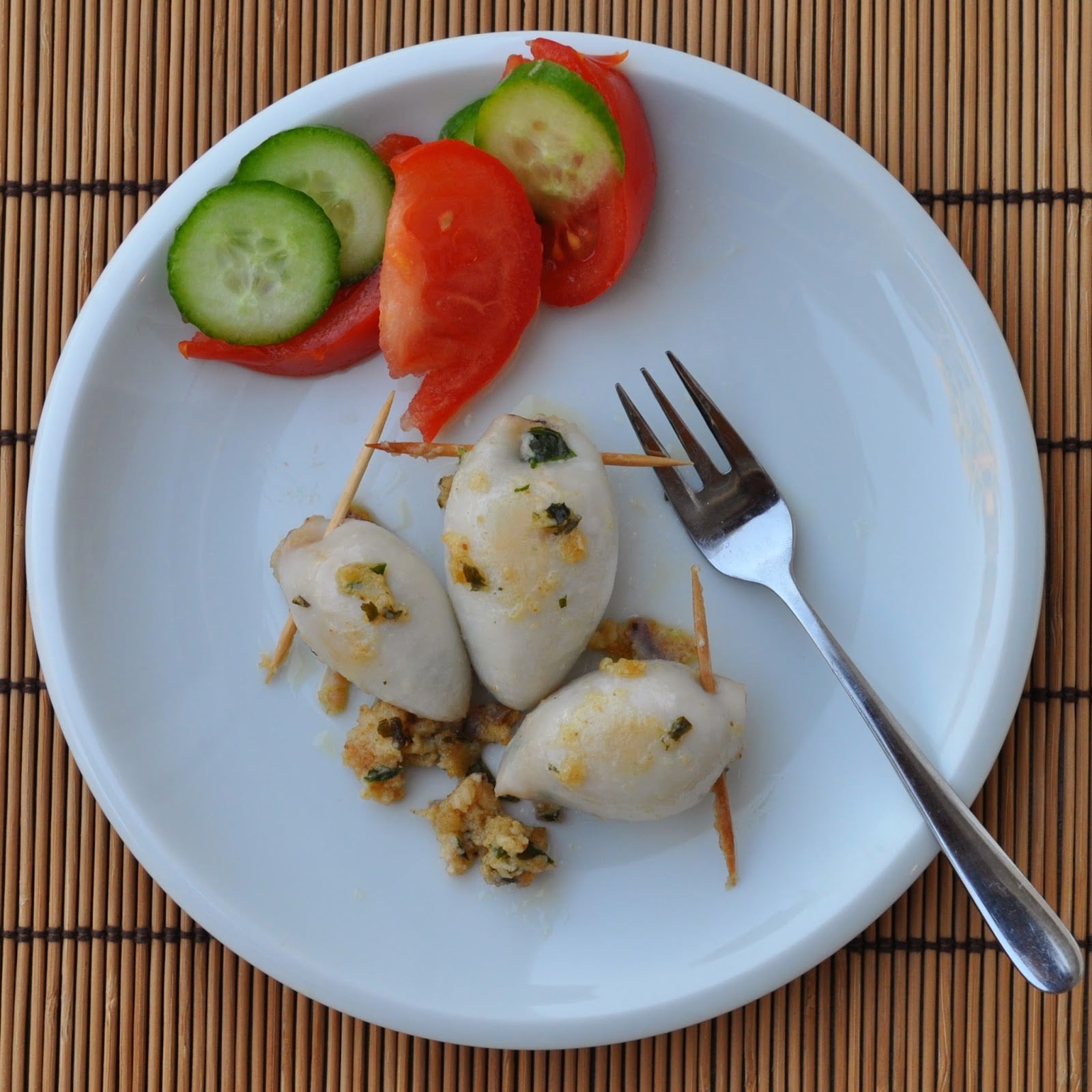 Three stuffed squid pieces secured with toothpicks on a white plate, garnished with cooked herbs. Slices of cucumber and tomato are placed on the side, with a small fork resting on the plate. The plate sits on a bamboo mat.