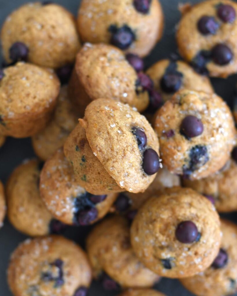 A close-up of a pile of mini blueberry muffins, topped with coarse sugar crystals and whole blueberries.