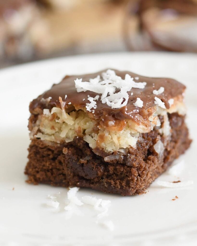 A close-up of a brownie topped with a layer of coconut and a thick layer of chocolate, garnished with shredded coconut on a white plate.