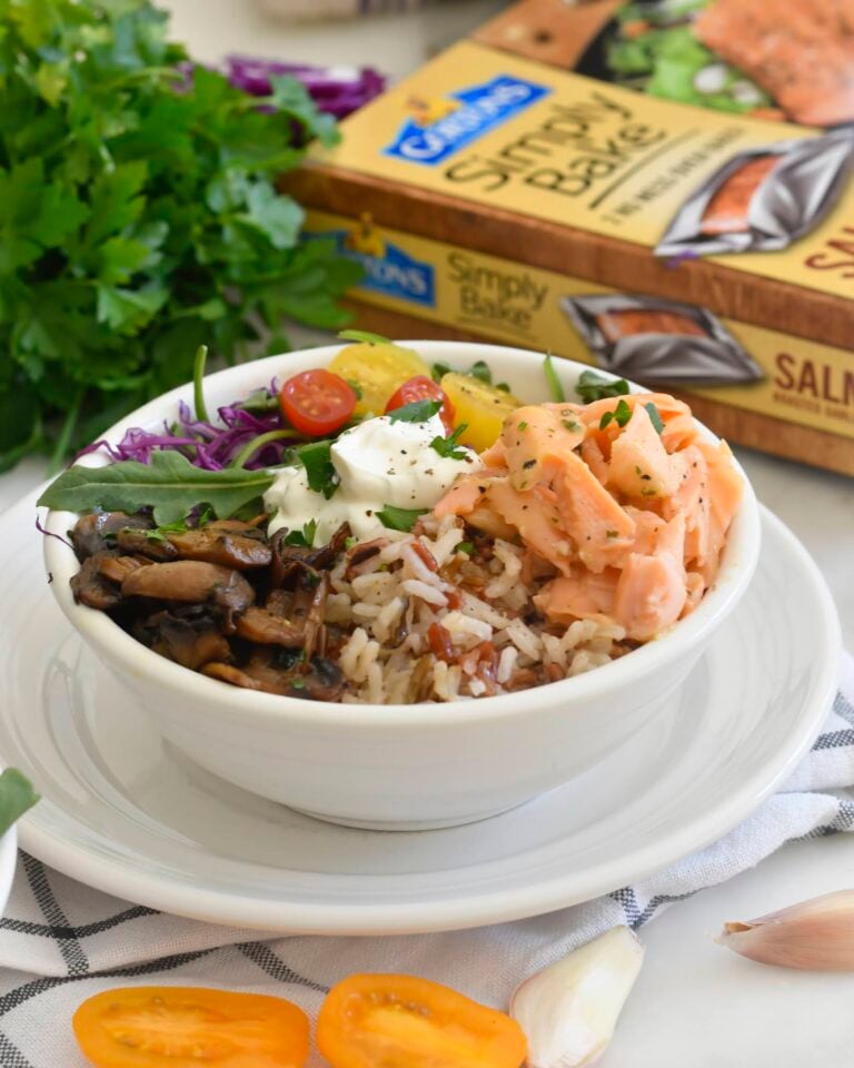 A bowl filled with rice, cooked salmon, mushrooms, cherry tomatoes, greens, purple cabbage, and a dollop of sauce, placed on a white plate with herbs, garlic, and a salmon bake package in the background.