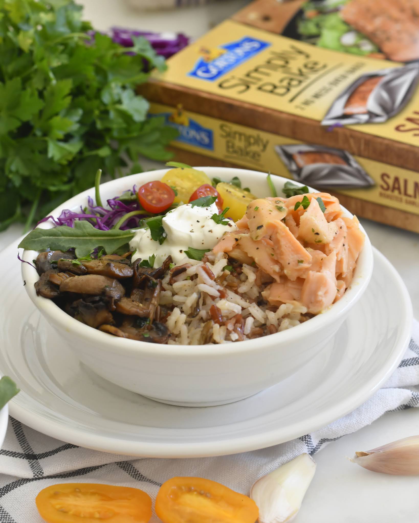 A bowl filled with rice, cooked salmon, mushrooms, cherry tomatoes, greens, purple cabbage, and a dollop of sauce, placed on a white plate with herbs, garlic, and a salmon bake package in the background.