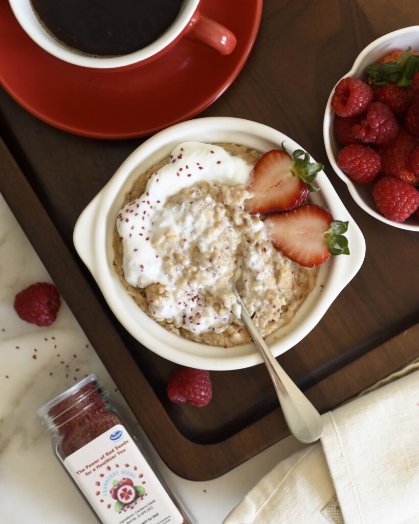 A bowl of oatmeal topped with yogurt, two sliced strawberries, and sprinkled seeds sits on a tray next to a cup of coffee, a bowl of fresh raspberries, and a jar of raspberry powder.