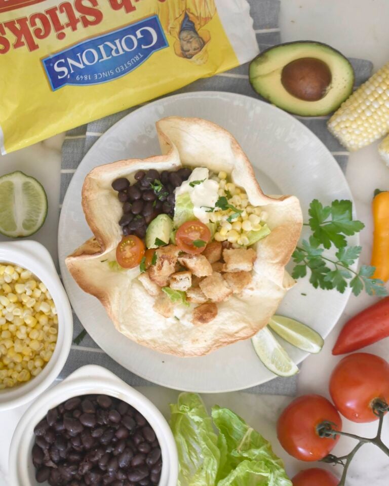 A tortilla bowl filled with black beans, lettuce, corn, cherry tomatoes, croutons, and cheese sits on a plate surrounded by avocado, corn, peppers, black beans, tomatoes, lettuce, and a bag of tortilla chips.