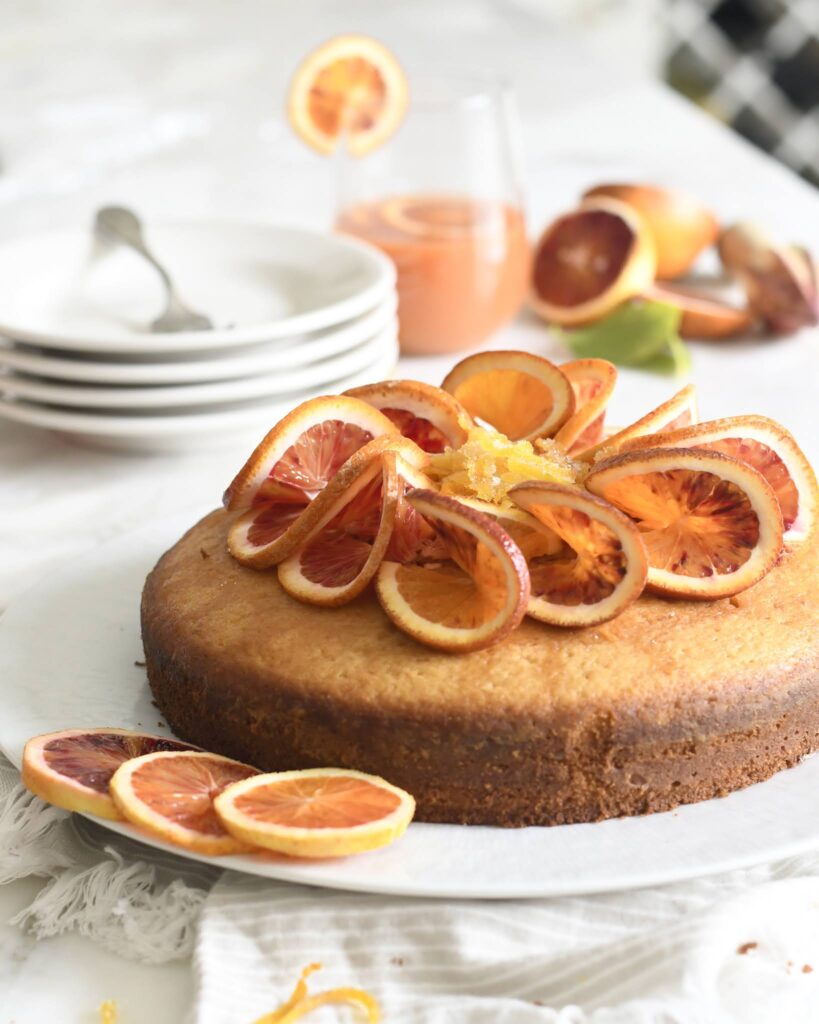 A round cake topped with fresh blood orange slices sits on a white plate. In the background, there are stacked plates, a glass of juice, and more blood oranges on a white tablecloth.