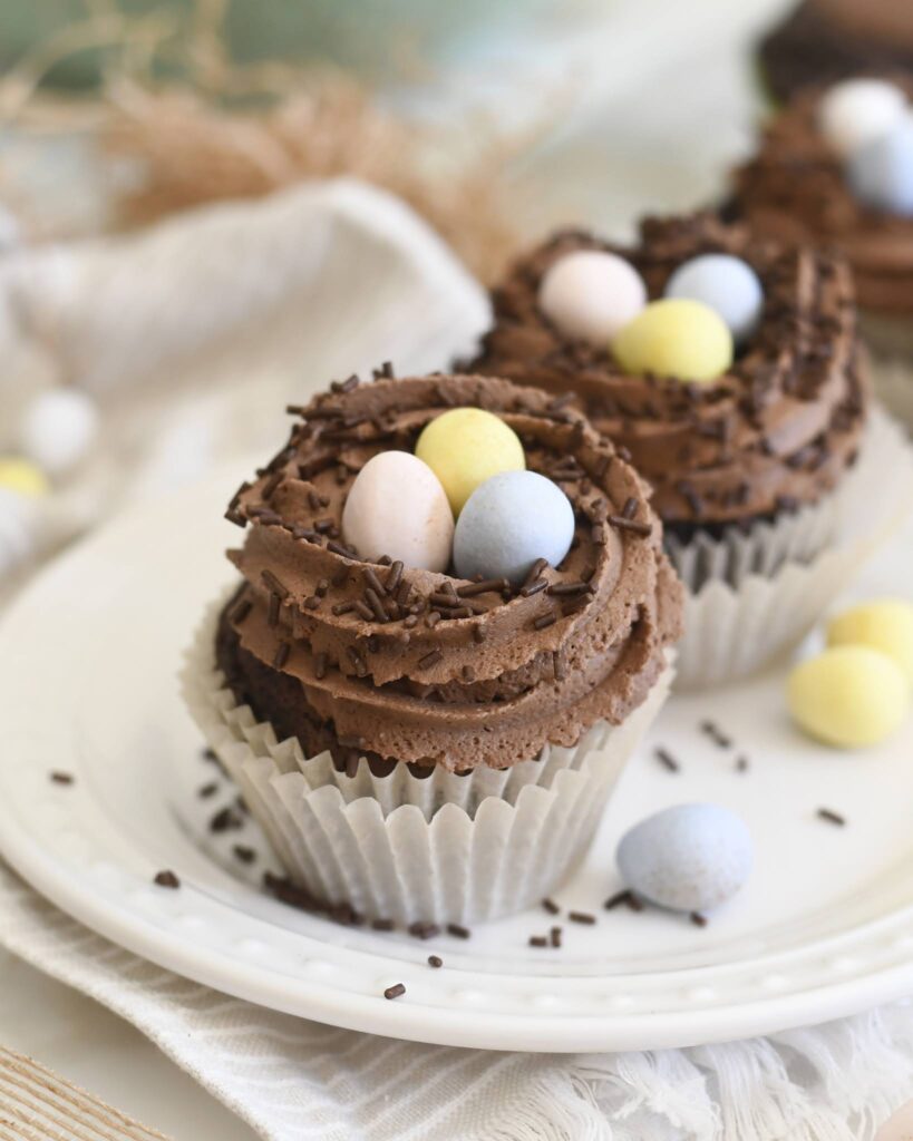 Chocolate cupcakes topped with swirls of chocolate frosting, chocolate sprinkles, and pastel-colored candy eggs, arranged on a white plate with a light-colored cloth underneath.