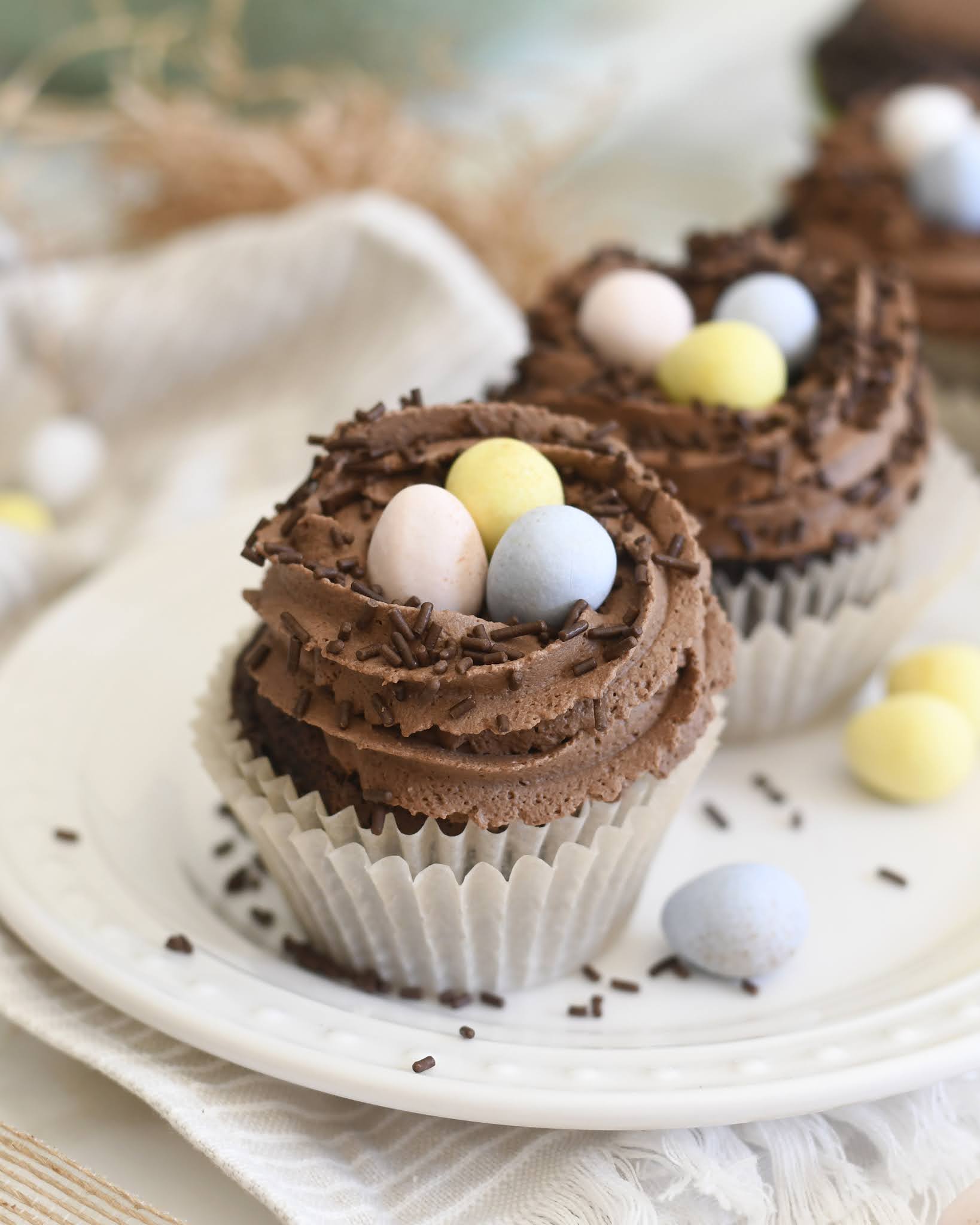 Chocolate cupcakes topped with swirls of chocolate frosting, chocolate sprinkles, and pastel-colored candy eggs, arranged on a white plate with a light-colored cloth underneath.